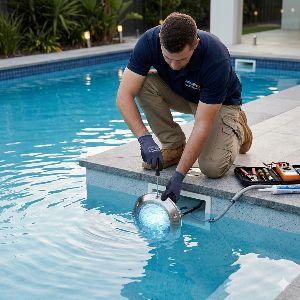Repair technician kneels at the pool edge, lowering a circular device into the water with a toolkit nearby.